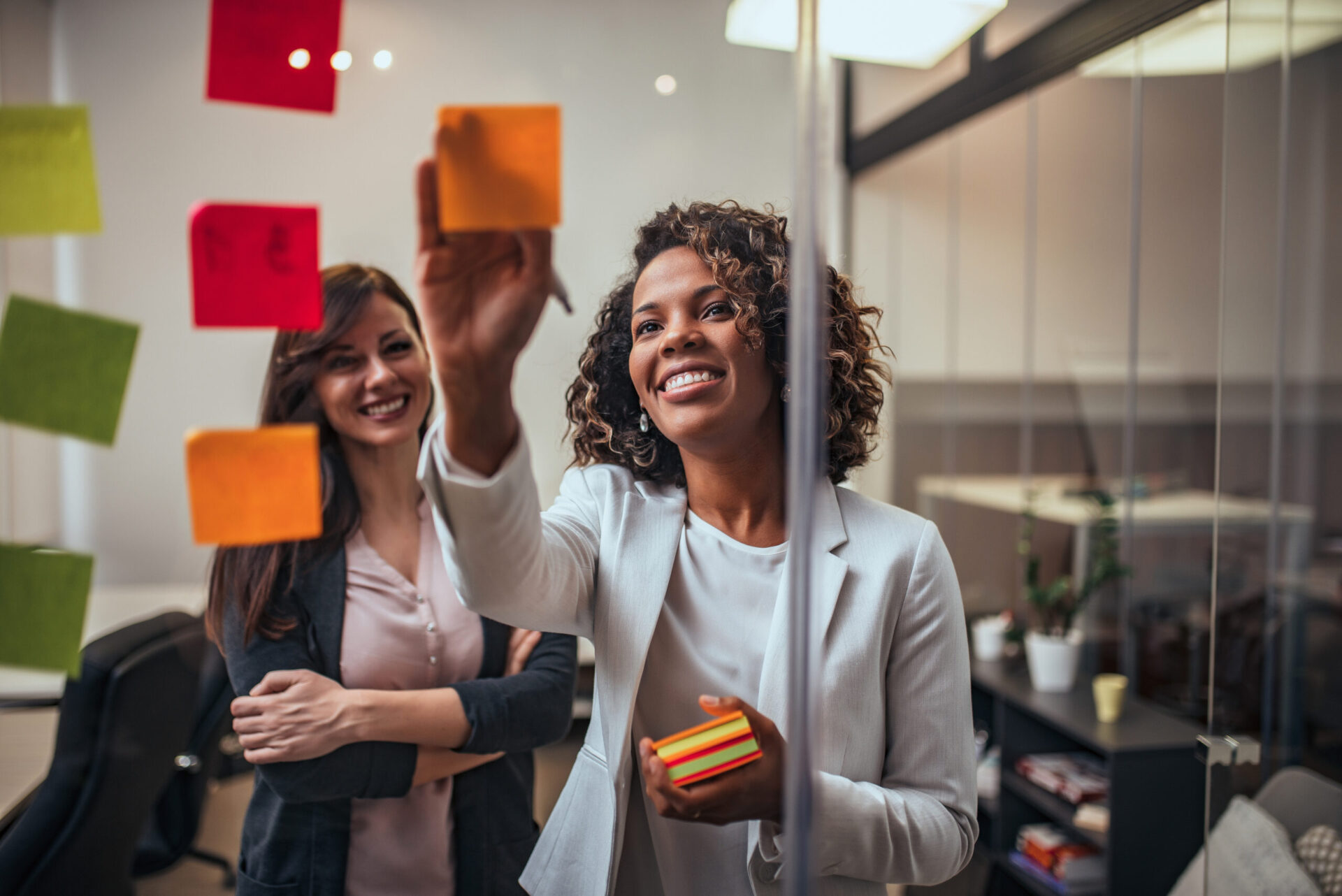 Creative businesswoman writing on sticky notes on a glass wall, female colleague looking. Creative businesswoman writing on sticky notes on a glass wall, female colleague looking.