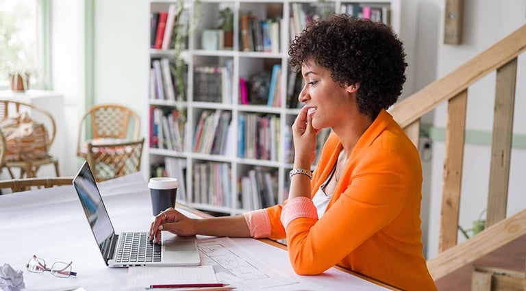 Black female professional working from home on a laptop with a coffee cup and glasses on her desk