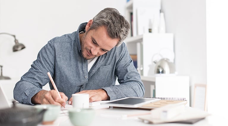 Adult man writing notes while sitting at a desk with notebooks and coffee cups