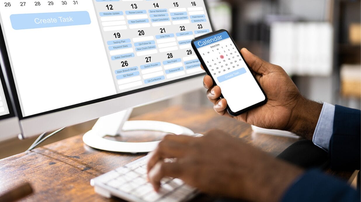Blog Covers – 2025-09-26T154906.629 Man holding a cell phone while sitting at his computer, reviewing calendar